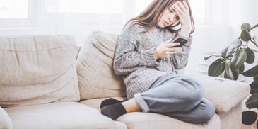 Young woman sits on couch looking alarmed at her smartphone