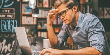 exhausted looking man working on a laptop in a room full of books