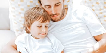 dad and son in white shirts lean on pillow while reading together