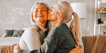 adult daughter with ponytail kisses smiling white haired mom 