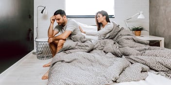 woman reaching for a man who is upset and turned away from her in bed