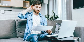 Man scratching the back of his head considering paperwork in front of his laptop