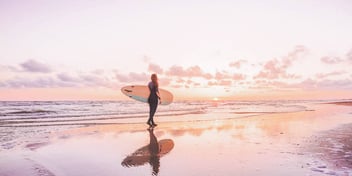 Woman walks with surfboard at sunset