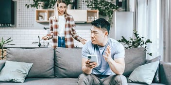 Young blond woman disagreeing with young Asian male partner that is sitting on a couch
